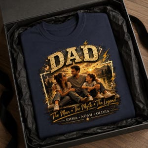 May include: Navy blue t-shirt with a graphic featuring the word "DAD" in a distressed gold font, a family portrait, and the text "The Man * The Myth * The Legend". The shirt is presented in a black gift box.