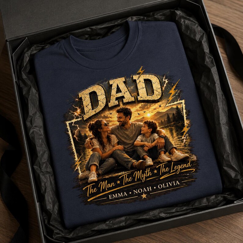 May include: Navy blue t-shirt with a graphic featuring the word "DAD" in a distressed gold font, a family portrait, and the text "The Man * The Myth * The Legend". The shirt is presented in a black gift box.