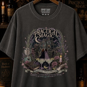 May include: A charcoal gray short-sleeve t-shirt featuring a graphic print of a house, two figures, a blender, two margaritas, and a book. The text reads Practical Magic, Midnight Margaritas, and There's a little witch in all of us.