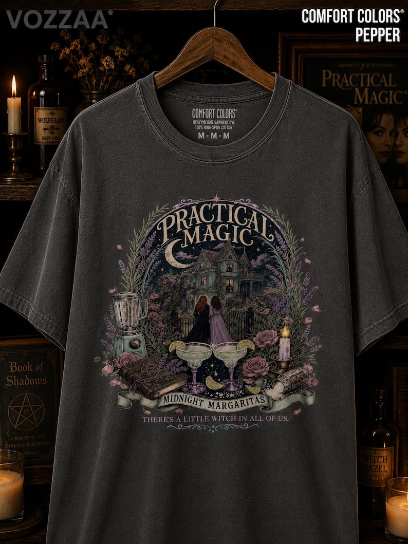 May include: A charcoal gray short-sleeve t-shirt featuring a graphic print of a house, two figures, a blender, two margaritas, and a book. The text reads Practical Magic, Midnight Margaritas, and There's a little witch in all of us.