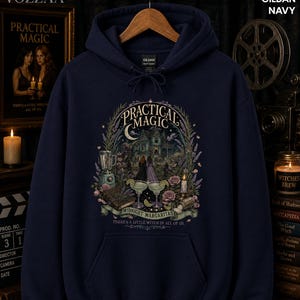 Op de afbeelding: Een marineblauwe hoodie met een grafische print met de tekst "Practical Magic" en "Midnight Margaritas" boven een illustratie van een huis, twee margarita's, een blender en bloemen. De tekst "There's a little witch in all of us" staat eronder.