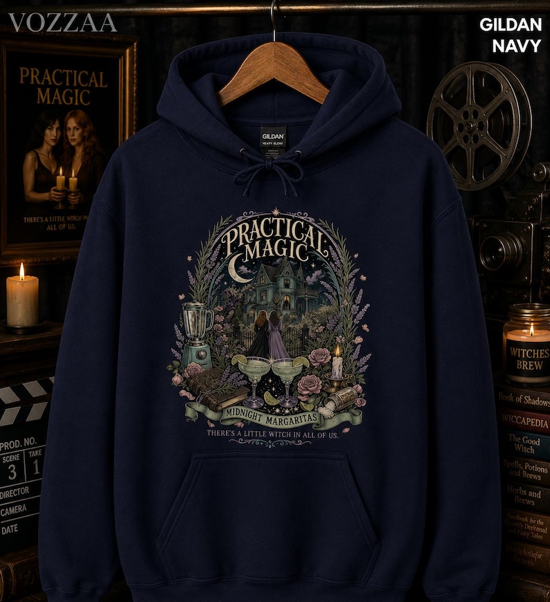 Op de afbeelding: Een marineblauwe hoodie met een grafische print met de tekst "Practical Magic" en "Midnight Margaritas" boven een illustratie van een huis, twee margarita's, een blender en bloemen. De tekst "There's a little witch in all of us" staat eronder.