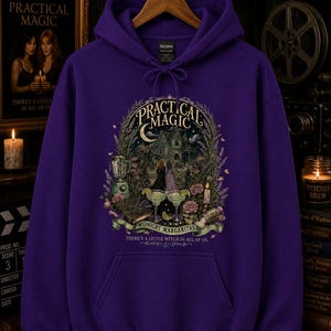 Op de afbeelding: Een paarse hoodie met een grafische print met de tekst Practical Magic, Midnight Margaritas en There's a little witch in all of us. De illustratie toont een huis, twee figuren, margarita's, bloemen en een blender.