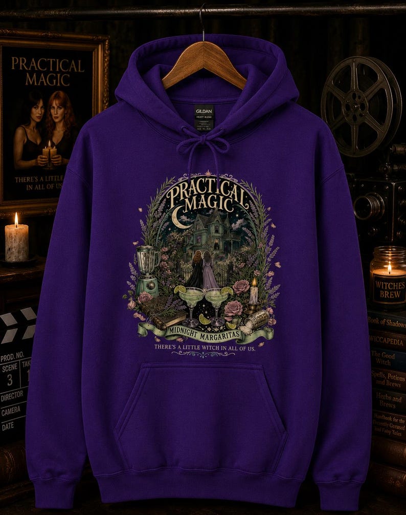 Op de afbeelding: Een paarse hoodie met een grafische print met de tekst Practical Magic, Midnight Margaritas en There's a little witch in all of us. De illustratie toont een huis, twee figuren, margarita's, bloemen en een blender.