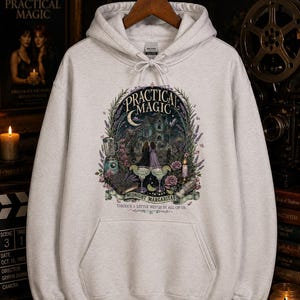 Op de afbeelding: Een lichtgrijze hoodie met een grafische print met de tekst "PRACTICAL MAGIC", "MIDNIGHT MARGARITAS" en "THERE'S A LITTLE WITCH IN ALL OF US". Het ontwerp bevat een huis, twee figuren, margarita's en bloemmotieven.