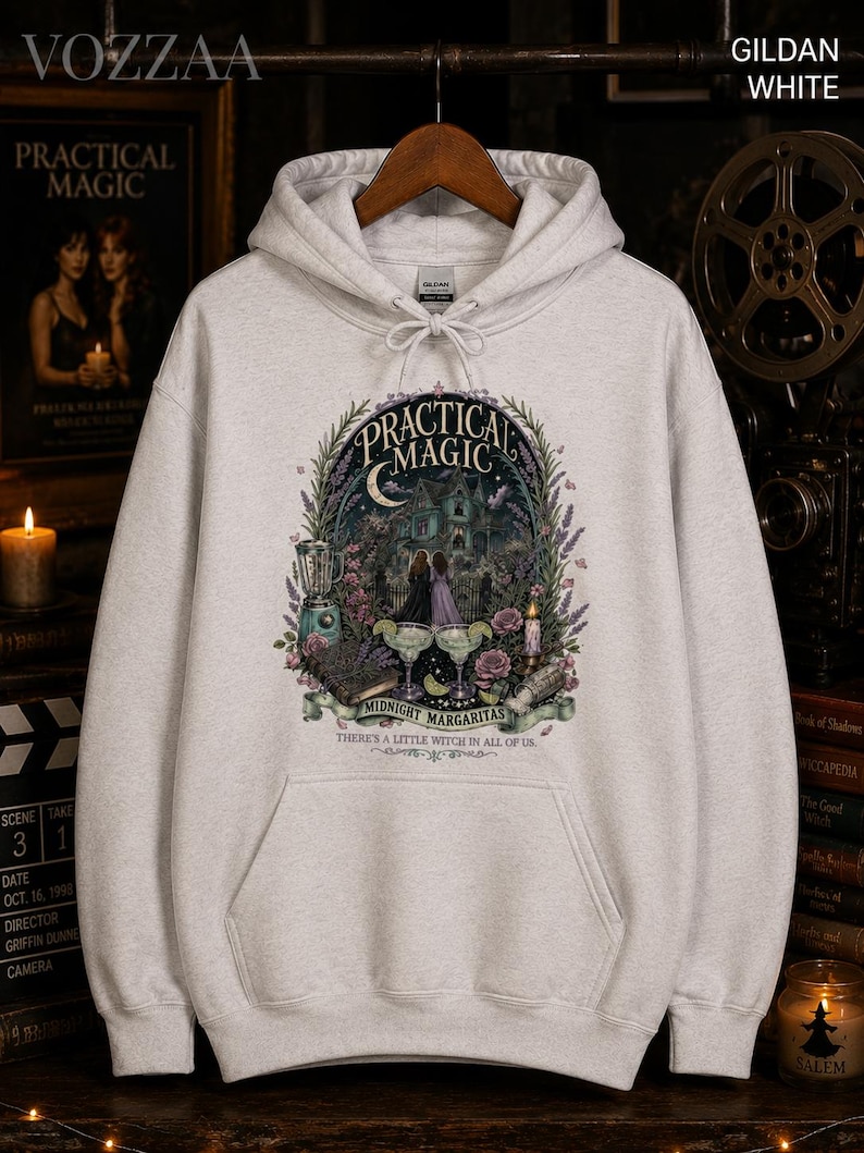 Op de afbeelding: Een lichtgrijze hoodie met een grafische print met de tekst "PRACTICAL MAGIC", "MIDNIGHT MARGARITAS" en "THERE'S A LITTLE WITCH IN ALL OF US". Het ontwerp bevat een huis, twee figuren, margarita's en bloemmotieven.