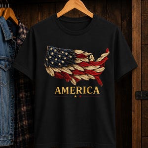 May include: A black short-sleeved t-shirt featuring a graphic of the United States map rendered in red, white, and blue feathers. The word AMERICA is printed in gold capital letters below the map, with three small stars underneath the text.