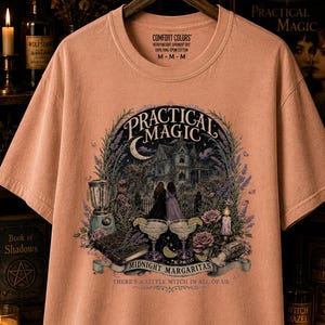 May include: A peachy-colored short-sleeve t-shirt featuring a graphic print of a house, two figures, margaritas, and a book. The text reads Practical Magic, Midnight Margaritas, and There's a little witch in all of us. The shirt has a crew neckline.