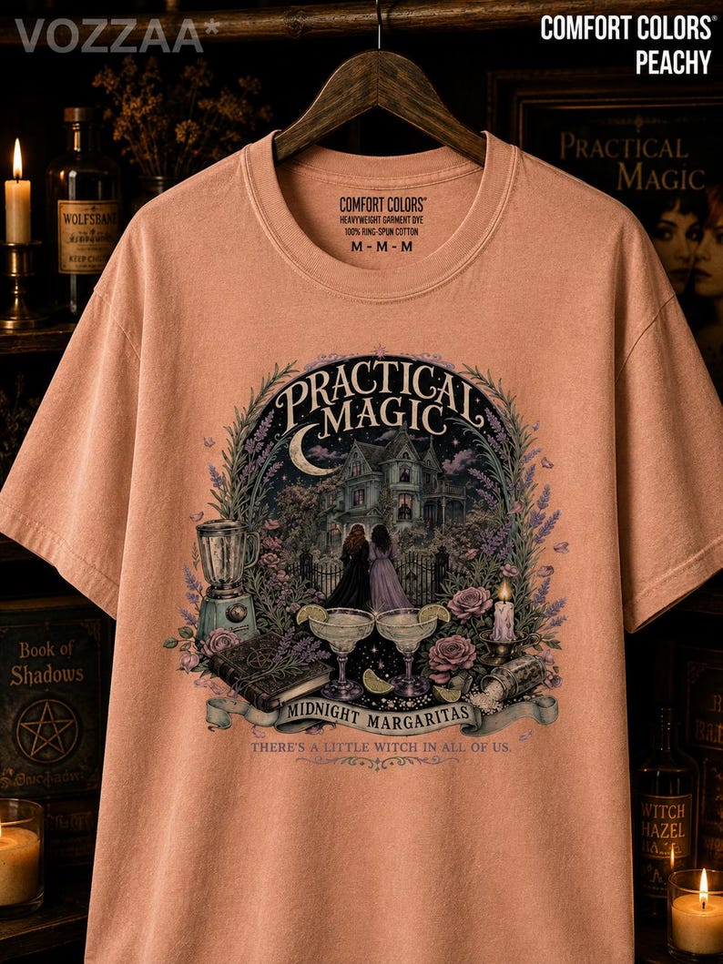 May include: A peachy-colored short-sleeve t-shirt featuring a graphic print of a house, two figures, margaritas, and a book. The text reads Practical Magic, Midnight Margaritas, and There's a little witch in all of us. The shirt has a crew neckline.