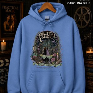 Op de afbeelding: Een Carolina blauwe hoodie met een grafische print met de tekst Practical Magic Midnight Margaritas There's a little witch in all of us. Het ontwerp bevat een huis, twee figuren, margarita's, een blender en bloemen.