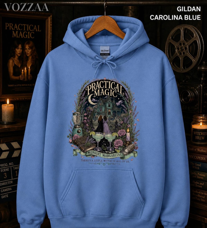 Op de afbeelding: Een Carolina blauwe hoodie met een grafische print met de tekst Practical Magic Midnight Margaritas There's a little witch in all of us. Het ontwerp bevat een huis, twee figuren, margarita's, een blender en bloemen.