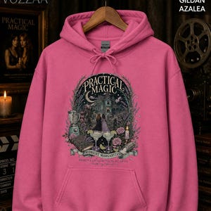 Op de afbeelding: Een felroze hoodie met een grafische print op de voorzijde. Het ontwerp toont een huis, twee figuren, een blender, twee margaritaglazen en een boek, met de tekst "PRACTICAL MAGIC MIDNIGHT MARGARITAS THERE'S A LITTLE WITCH IN ALL OF US."