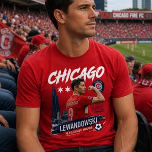 May include: A red short-sleeved t-shirt featuring a graphic print of a soccer player flexing, the Chicago skyline, the text CHICAGO, LEWANDOWSKI, THE POLISH GOAL MACHINE, a soccer ball, two stars, and a circular team logo on the front chest area.