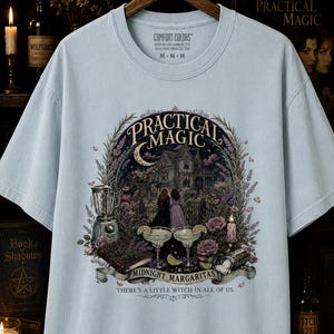 May include: A light blue short-sleeve t-shirt featuring a graphic print of a Victorian house, two figures, a blender, two margaritas, and a book. The text reads Practical Magic, Midnight Margaritas, and There's a little witch in all of us.