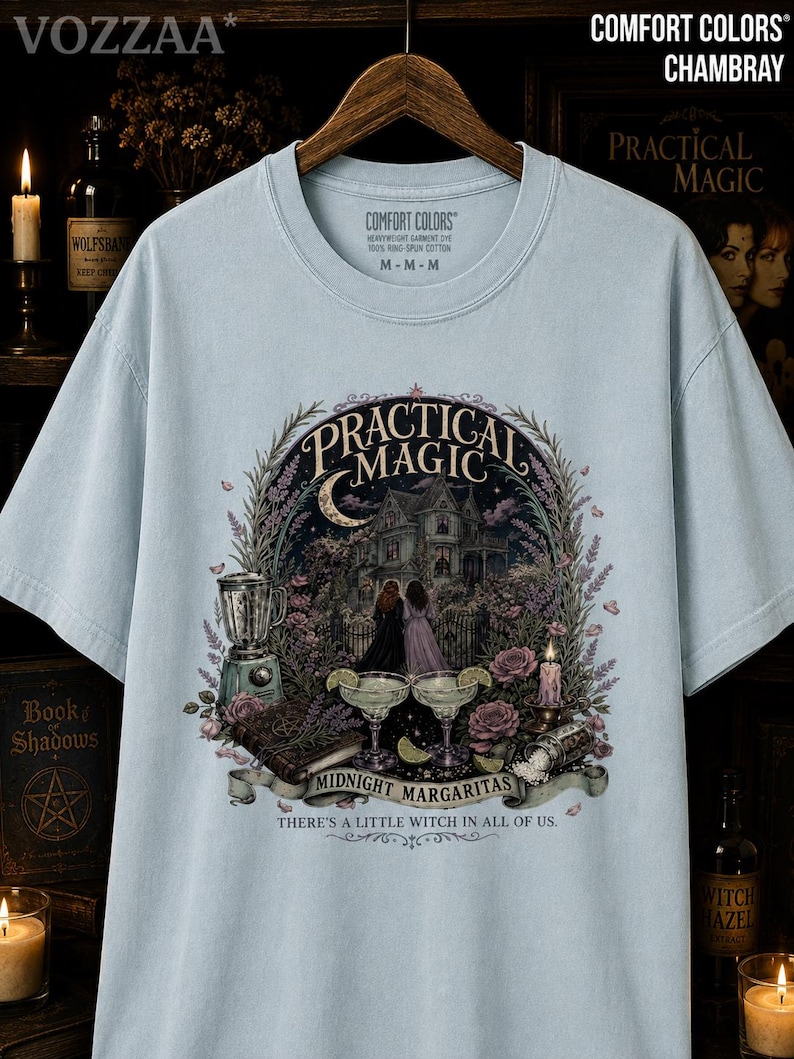 May include: A light blue short-sleeve t-shirt featuring a graphic print of a Victorian house, two figures, a blender, two margaritas, and a book. The text reads Practical Magic, Midnight Margaritas, and There's a little witch in all of us.