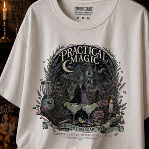 May include: A white t-shirt featuring a graphic print with the text "PRACTICAL MAGIC", "MIDNIGHT MARGARITAS", and "THERE'S A LITTLE WITCH IN ALL OF US." The design includes a house, two figures, a blender, two margaritas, a book, and flowers.