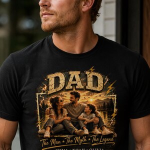 May include: Black t-shirt featuring a graphic with the word "DAD" in a distressed gold font. The graphic includes a family portrait with the text "The Man * The Myth * The Legend" and the names Emma, Noah, and Olivia.