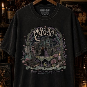 May include: A black short-sleeve t-shirt featuring a graphic print with the text "PRACTICAL MAGIC", "MIDNIGHT MARGARITAS", and "THERE'S A LITTLE WITCH IN ALL OF US". The design includes a house, two people, margaritas, a blender, and flowers.