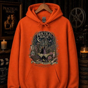 Op de afbeelding: Een oranje hoodie met een grafische print met de tekst Practical Magic, Midnight Margaritas en There's a Little Witch in All of Us. De illustratie toont een huis, twee figuren, een blender en twee margaritaglazen.