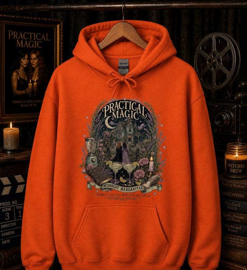 Op de afbeelding: Een oranje hoodie met een grafische print met de tekst Practical Magic, Midnight Margaritas en There's a Little Witch in All of Us. De illustratie toont een huis, twee figuren, een blender en twee margaritaglazen.