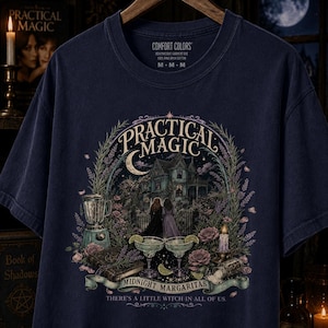 May include: A navy blue t-shirt featuring a graphic print with the text "Practical Magic" and "Midnight Margaritas" along with "There's a little witch in all of us." The design includes a house, two people, a blender, margaritas, and flowers.