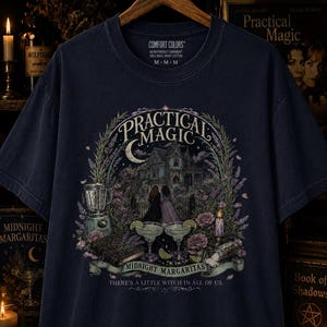 May include: A dark navy blue t-shirt featuring a graphic print of a Victorian house, two figures, a blender, two margaritas, and roses. The text reads Practical Magic, Midnight Margaritas, and There's a little witch in all of us. The shirt is size M.