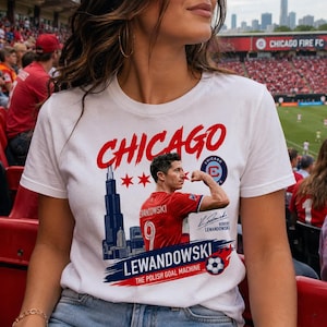 May include: A white t-shirt featuring a graphic of Robert Lewandowski flexing his arm, the Chicago skyline, and the text "CHICAGO", "LEWANDOWSKI", "THE POLISH GOAL MACHINE", and a signature. The design includes red stars, a soccer ball, and a logo.