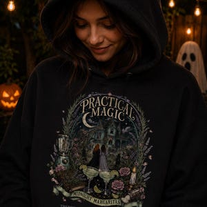 Op de afbeelding: Een zwarte hoodie met een grafische print met de tekst "PRACTICAL MAGIC", "MIDNIGHT MARGARITAS" en "THERE'S A LITTLE WITCH IN ALL OF US." Het ontwerp bevat een huis, twee figuren, een blender en twee margaritaglazen.