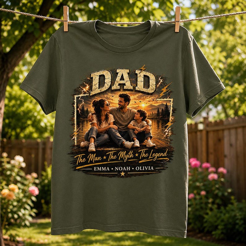 May include: Olive green t-shirt with a graphic featuring the word "DAD" in a distressed font above a family portrait. The text "The Man * The Myth * The Legend" and names are below the image. The shirt is hanging on a clothesline.