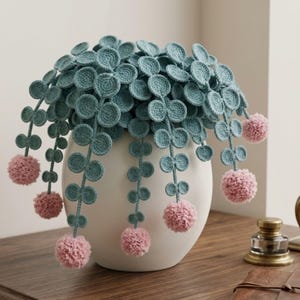 May include: A decorative plant sculpture featuring crocheted teal circular leaves on trailing stems, accented with soft pink pom-poms at the ends. The plant is housed in a smooth, rounded white ceramic vase, creating a textured and layered appearance.