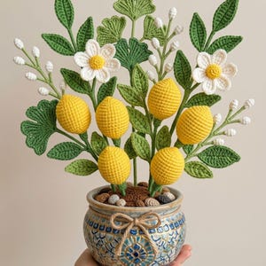 Puede incluir: Un limonero tejido a mano en un macetero de cerámica decorativo. El árbol presenta limones amarillos, flores blancas y hojas verdes. La maceta es azul y blanca con un detalle de lazo.