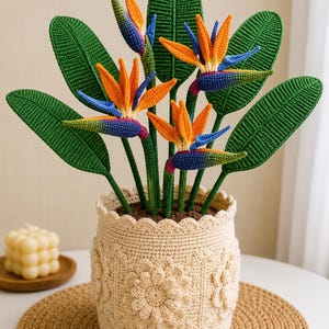 Puede incluir: Una planta de ganchillo de Ave del Paraíso en una maceta beige. Las flores son naranjas, azules y moradas, con hojas verdes. La maceta tiene un borde festoneado y diseños florales. La planta se exhibe sobre una estera redonda tejida.