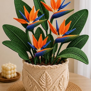 Könnte beinhalten: Eine gehäkelte Paradiesvogelblume mit leuchtend orangefarbenen und blauen Blütenblättern sowie strukturierten grünen Blättern, in einem beigen gehäkelten Korb mit einem erhabenen Blattmuster an der Außenseite. Die Pflanze hat mehrere Stängel.