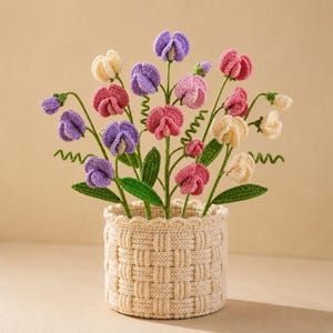 May include: A handcrafted crochet floral arrangement featuring colorful sweet pea flowers in shades of pink, purple, and cream. The flowers are arranged in a woven basket, creating a charming and decorative piece.