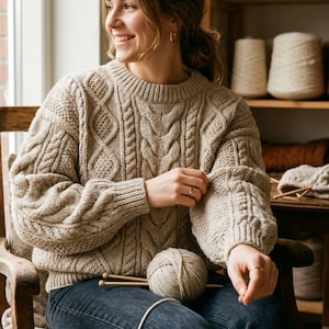 May include: A beige cable-knit sweater with a round neckline and long sleeves. The sweater features a textured pattern and is paired with blue jeans. A ball of yarn and knitting needles are visible.