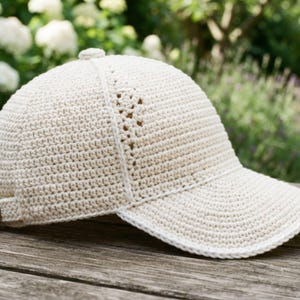 Puede incluir: Una gorra de béisbol de color crema hecha de hilo de ganchillo. La gorra presenta una corona estructurada con un pequeño botón en la parte superior, una visera curva y un patrón decorativo de punto abierto que recorre los paneles laterales.