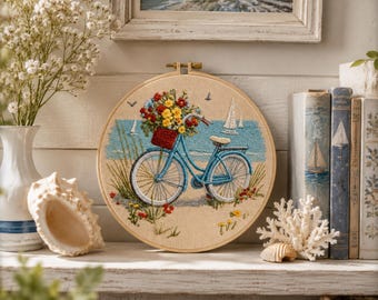 Puede incluir: Un bastidor de bordado circular que presenta una escena cosida de una bicicleta azul claro con una cesta roja llena de flores amarillas y rojas. El fondo representa una playa de arena, olas azules, hierba alta y dos veleros blancos.