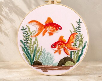 May include: A circular wooden embroidery hoop containing a fabric panel with hand-stitched thread art. The design features two bright orange goldfish swimming among various green aquatic plants and brown rocks on a pale pink background.