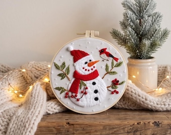 Puede incluir: Un bastidor de bordado circular con fondo de tela blanca que muestra un muñeco de nieve bordado con gorro y bufanda rojos, nariz naranja y botones negros. El diseño incluye ramas de pino verdes, bayas rojas y un pequeño pájaro cardenal rojo.