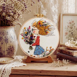 May include: A circular embroidery hoop featuring a stitched illustration of a person with windblown hair holding a large bouquet of colorful wildflowers. The person wears a red long-sleeved top and a blue skirt, with small flowers at their feet.