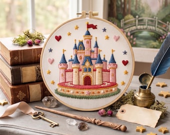 Könnte beinhalten: Ein runder Stickrahmen mit einem handgestickten Märchenschloss mit rosafarbenen Türmen, blauen kegelförmigen Dächern und einem roten Torbogen. Das Design ist mit gelben Sternen, roten und rosafarbenen Herzen sowie einer grünen Grasbasis verziert.