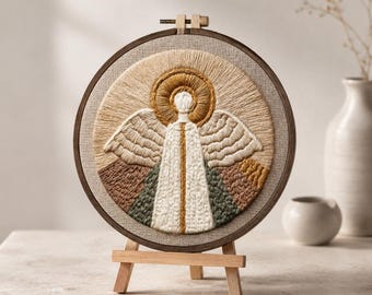 Puede incluir: Un bastidor de bordado circular que contiene una pieza de arte textil con un ángel estilizado con alas blancas, un halo dorado y una túnica blanca larga con detalles verticales dorados y verdes, exhibido en un pequeño caballete de madera.