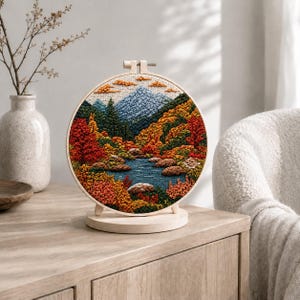 May include: A circular embroidery hoop art piece featuring a landscape with a blue mountain, a central stream, and surrounding autumn foliage in shades of red, orange, yellow, and green. The embroidery is mounted on a light wood circular stand.