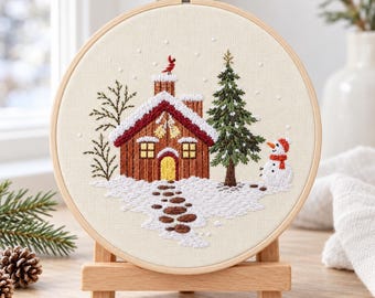 Puede incluir: Un bastidor de bordado circular con un panel de tela que muestra una escena invernal. El diseño presenta una cabaña marrón con techo nevado, un pino verde, un muñeco de nieve con bufanda naranja y un pájaro rojo en la chimenea.