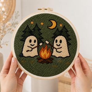 Peut inclure: Un tambour à broder circulaire en punch needle représentant deux fantômes beiges aux yeux et bouches noirs, tenant chacun un bâton au-dessus d'un feu de camp orange et jaune. Le fond est vert foncé avec des sapins et des étoiles jaunes.