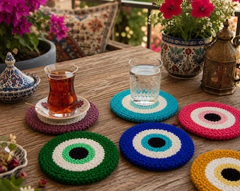 Puede incluir: Una colección de posavasos redondos de ganchillo con un patrón concéntrico en forma de ojo en varios colores, incluyendo verde, azul, rosa y amarillo. Cada posavasos tiene un círculo negro central rodeado de blanco y un color exterior.