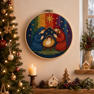 Può includere: Un'opera d'arte circolare su telaio da ricamo che raffigura una scena della natività con una figura in blu, una figura in rosso e un bambino in una mangiatoia. Lo sfondo presenta un gradiente arcobaleno con stelle gialle e una grande stella.