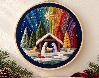 Puede incluir: Un bastidor de bordado circular que muestra una escena de nacimiento bajo un cielo de colores arcoíris que recuerda a una aurora boreal. Incluye pinos bordados, un establo de madera, figuras y animales pequeños.