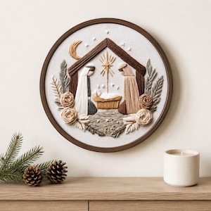Puede incluir: Una pieza de arte en bastidor de bordado circular que presenta una escena de natividad cosida con hilo sobre tela blanca. El diseño incluye dos figuras con túnicas, un pesebre, una estrella y una luna creciente, enmarcado por follaje y flores.