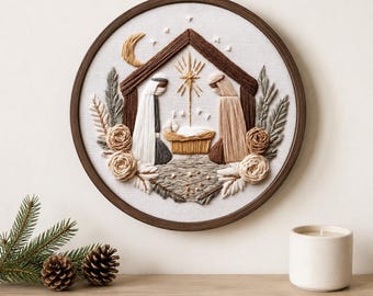 Puede incluir: Una pieza de arte en bastidor de bordado circular que presenta una escena de natividad cosida con hilo sobre tela blanca. El diseño incluye dos figuras con túnicas, un pesebre, una estrella y una luna creciente, enmarcado por follaje y flores.
